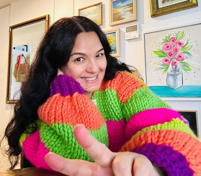 May include: A person wearing a chunky knit sweater with vibrant horizontal stripes of orange, green, pink, and purple. The sweater has a wide, oversized fit. The person is smiling and giving a peace sign.