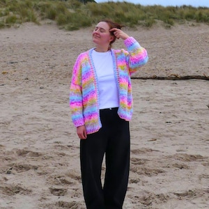 May include: A rainbow-striped cardigan in pink, yellow, blue, and purple. The open-front sweater is worn with a white t-shirt, black trousers, and blue shoes. The cardigan is a striking, colourful garment.