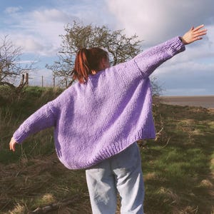 May include: A lilac-colored, oversized knit sweater with long sleeves. The sweater is worn with light blue jeans. The person is standing outdoors with arms outstretched, against a backdrop of a cloudy sky and grassy field.