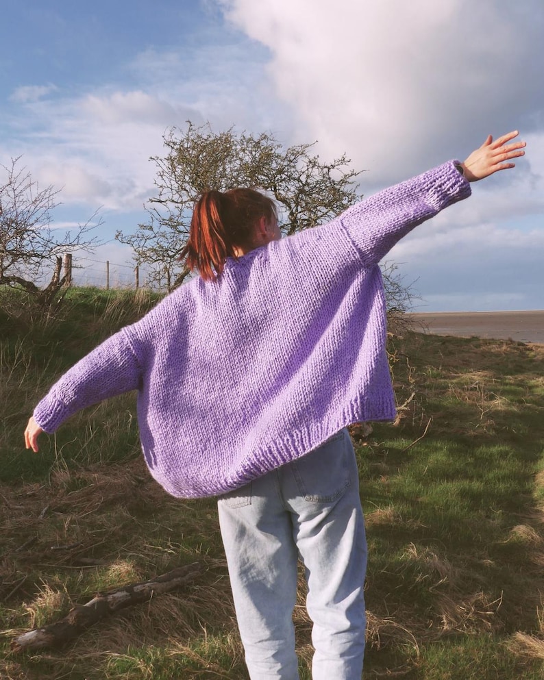 May include: A lilac-colored, oversized knit sweater with long sleeves. The sweater is worn with light blue jeans. The person is standing outdoors with arms outstretched, against a backdrop of a cloudy sky and grassy field.