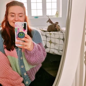 May include: A colorful, chunky knit cardigan in shades of pink, green, blue, and purple. The sweater has large, puffy sleeves and is buttoned up the front with decorative buttons. The person in the image is holding a phone with a floral case.
