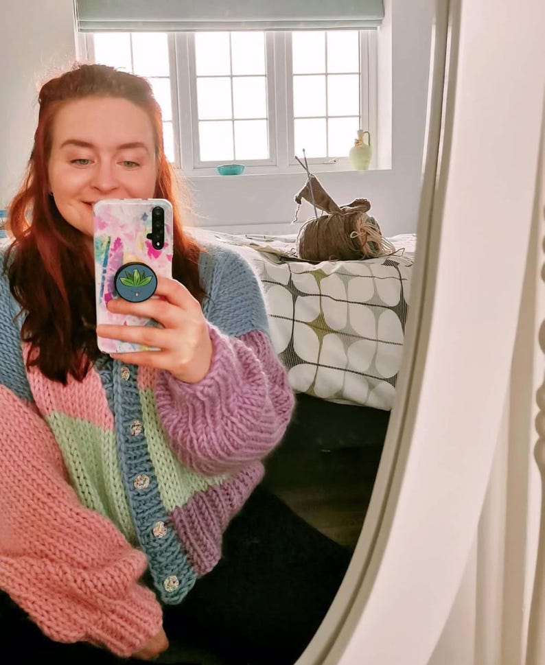 May include: A colorful, chunky knit cardigan in shades of pink, green, blue, and purple. The sweater has large, puffy sleeves and is buttoned up the front with decorative buttons. The person in the image is holding a phone with a floral case.