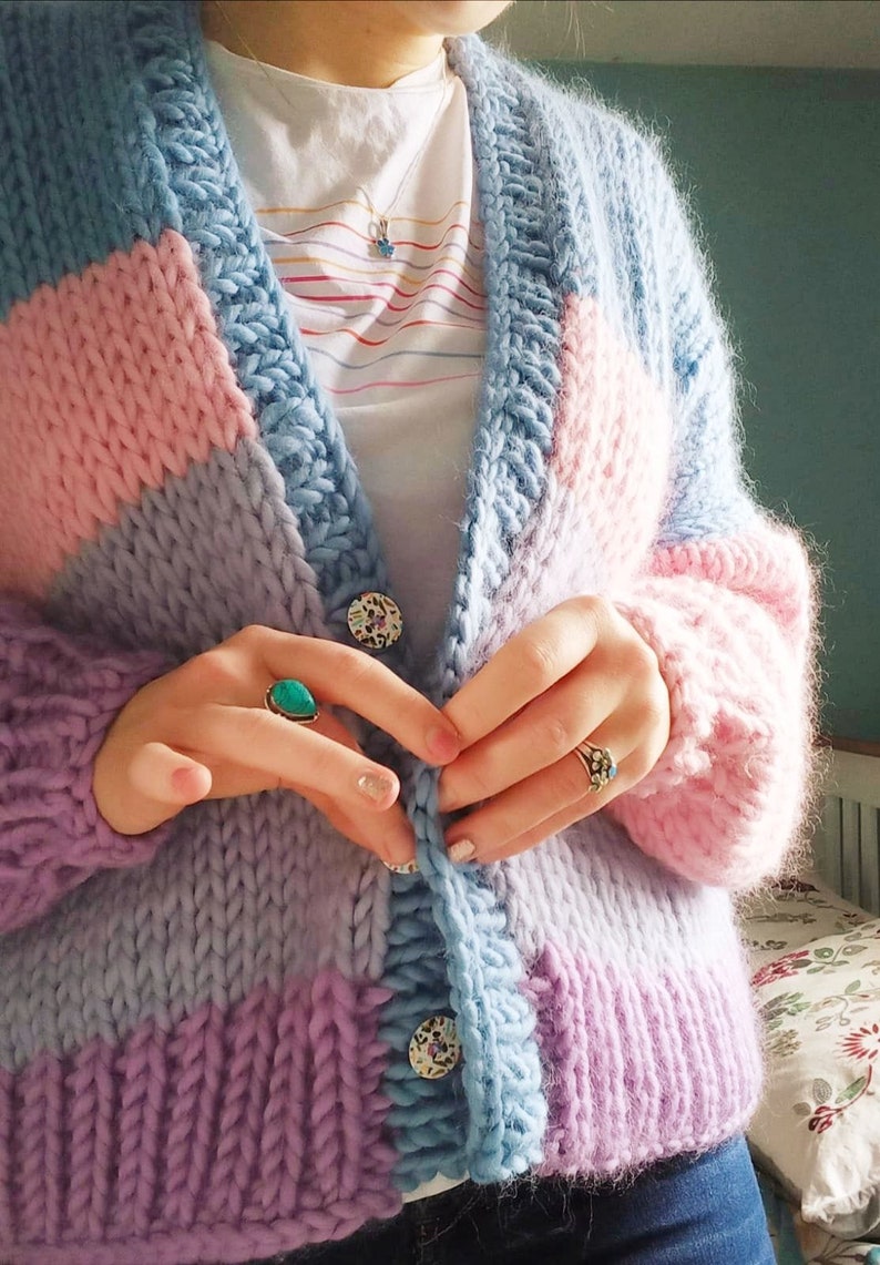May include: A chunky knit cardigan in pastel colors: light blue, pink, and lavender. The oversized sweater has large buttons and a thick, textured knit. The cardigan is worn over a white t-shirt.