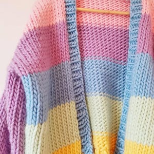 May include: A chunky knit cardigan in a patchwork design. The sweater features blocks of pastel colors including pink, purple, blue, yellow, and green. The cardigan is hanging on a wooden hanger.