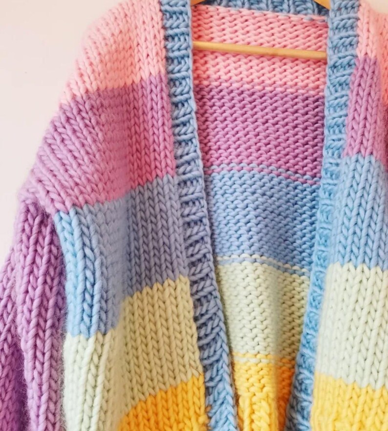 May include: A chunky knit cardigan in a patchwork design. The sweater features blocks of pastel colors including pink, purple, blue, yellow, and green. The cardigan is hanging on a wooden hanger.