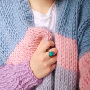 May include: A chunky knit cardigan in pastel colors. The sweater features a color block design with light blue, pink, and lavender sections. A gold ring with a turquoise stone is visible. The person is wearing a white t-shirt and a silver necklace.