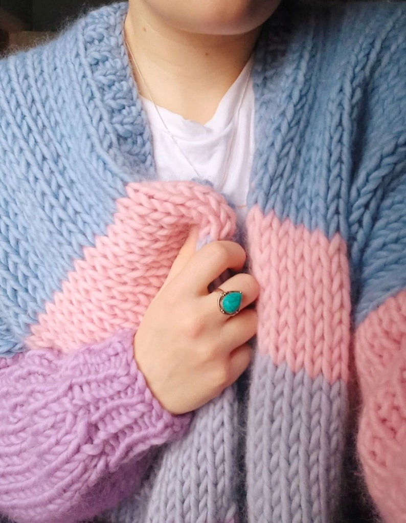 May include: A chunky knit cardigan in pastel colors. The sweater features a color block design with light blue, pink, and lavender sections. A gold ring with a turquoise stone is visible. The person is wearing a white t-shirt and a silver necklace.