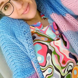 May include: A light blue, chunky knit cardigan with a matching blue knit hood. The cardigan is layered over a colorful patterned top with pink, green, and blue swirls. The outfit is a mix of textures and colors.