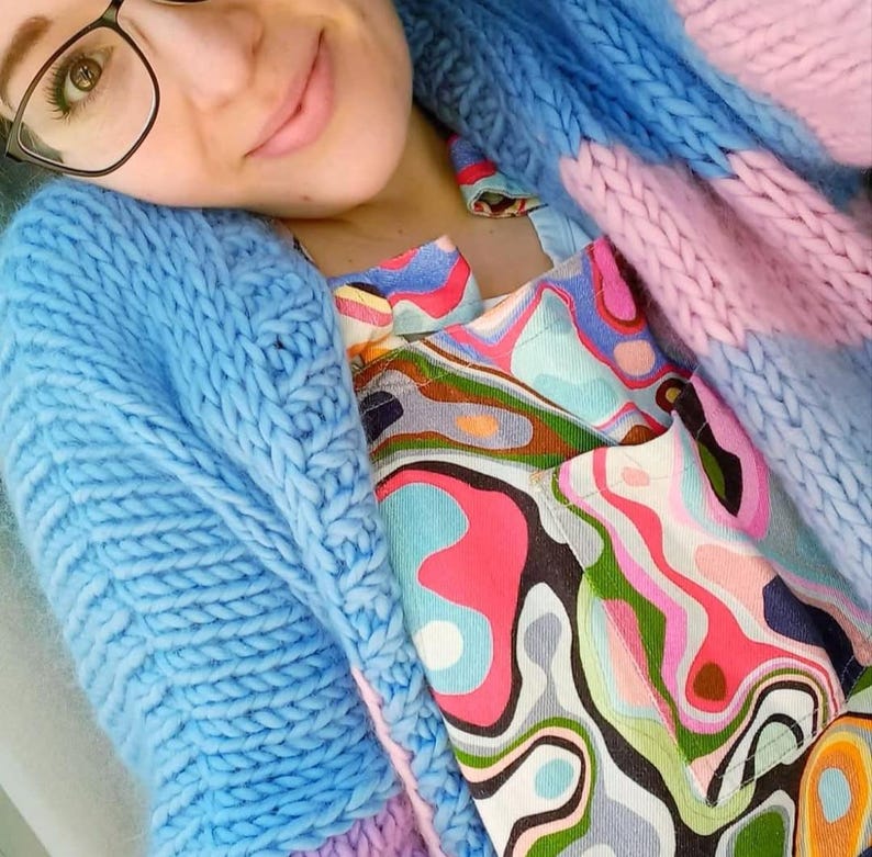 May include: A light blue, chunky knit cardigan with a matching blue knit hood. The cardigan is layered over a colorful patterned top with pink, green, and blue swirls. The outfit is a mix of textures and colors.