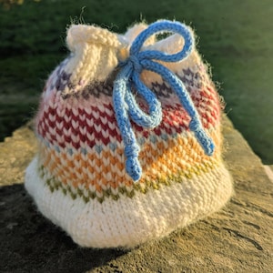 May include: A hand-knitted drawstring bag with a white base and a colorful striped pattern in red, orange, green, and purple. The bag is cinched with a blue cord. The bag is sitting on a stone surface.