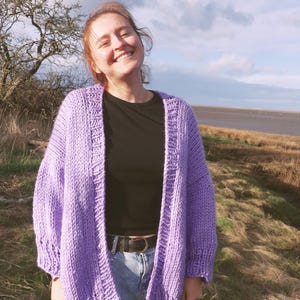 May include: A lilac-colored, chunky knit cardigan with a relaxed fit. The open-front sweater features a wide ribbed hem and cuffs. The model is wearing a black top and blue jeans. The background includes a grassy field, a tree, and a cloudy sky.