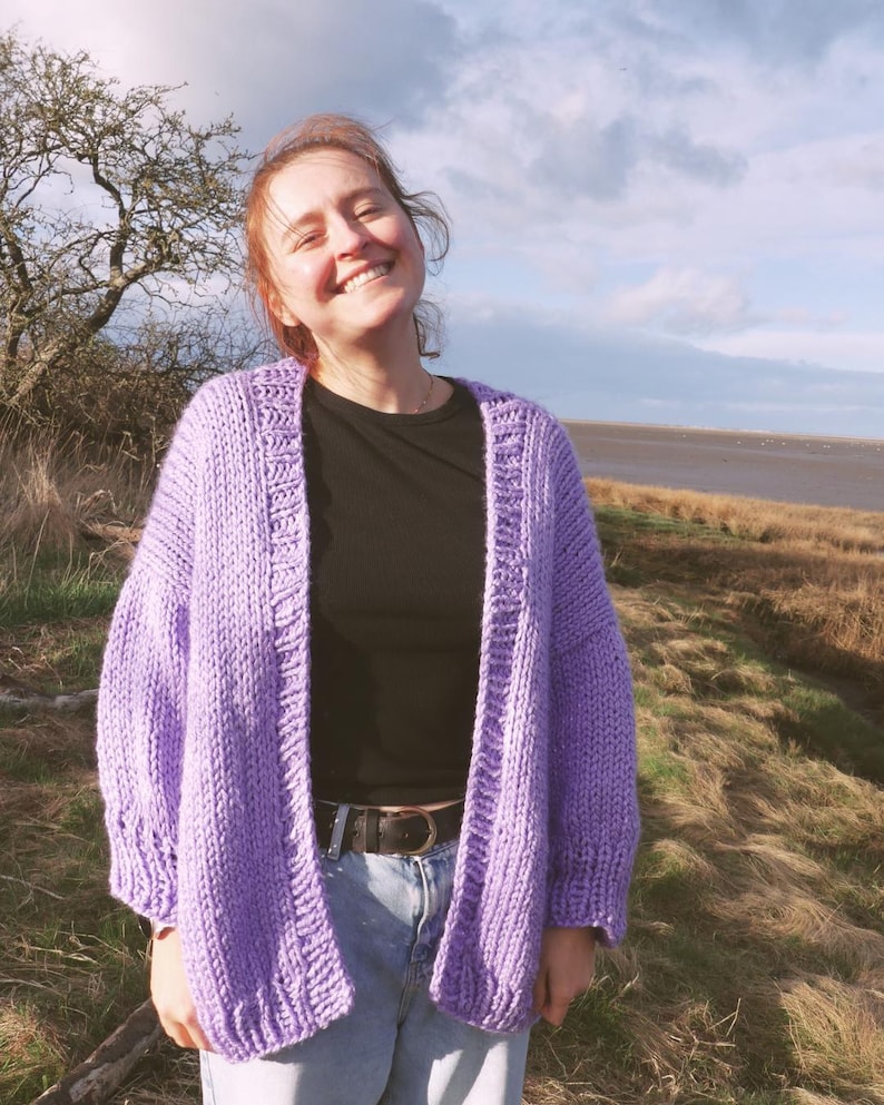 May include: A lilac-colored, chunky knit cardigan with a relaxed fit. The open-front sweater features a wide ribbed hem and cuffs. The model is wearing a black top and blue jeans. The background includes a grassy field, a tree, and a cloudy sky.