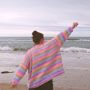 May include: A vibrant, striped sweater in pastel shades of pink, yellow, blue, and green. The sweater features long sleeves and a loose fit. The person is on a sandy beach with the sea in the background.