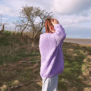 May include: A lilac-colored, chunky knit sweater with long sleeves. The sweater is worn with light-wash jeans. The background features a grassy field, trees, and a cloudy sky.
