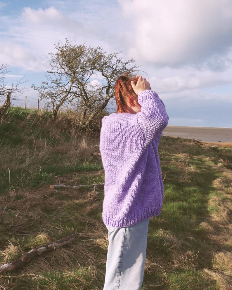 May include: A lilac-colored, chunky knit sweater with long sleeves. The sweater is worn with light-wash jeans. The background features a grassy field, trees, and a cloudy sky.