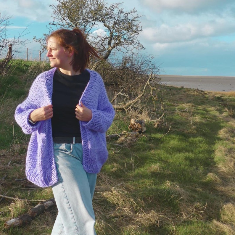 May include: A woman wearing a light purple, chunky knit cardigan over a black top and blue jeans. The cardigan is open, revealing the outfit underneath. The background features a grassy area, trees, and a cloudy sky.