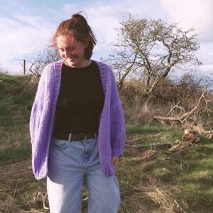 May include: A lilac-colored, open-front cardigan with a chunky knit design. The cardigan is worn over a black top and light blue jeans. The outfit is completed with a black belt. The background features a grassy field and a tree under a partly cloudy sky.