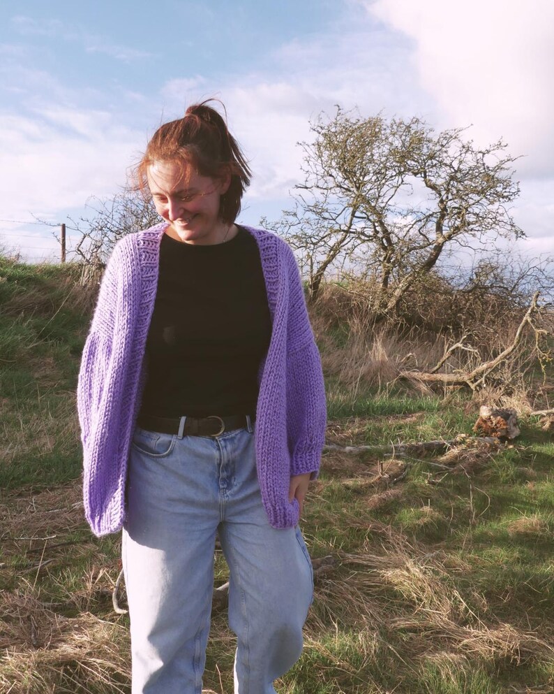 May include: A lilac-colored, open-front cardigan with a chunky knit design. The cardigan is worn over a black top and light blue jeans. The outfit is completed with a black belt. The background features a grassy field and a tree under a partly cloudy sky.