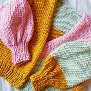 May include: A close-up view of a hand-knitted sweater in shades of pink, mustard yellow, and mint green. The sweater features large, puffy sleeves and a cozy, textured knit design. The color-blocked pattern adds a modern touch to the handmade garment.