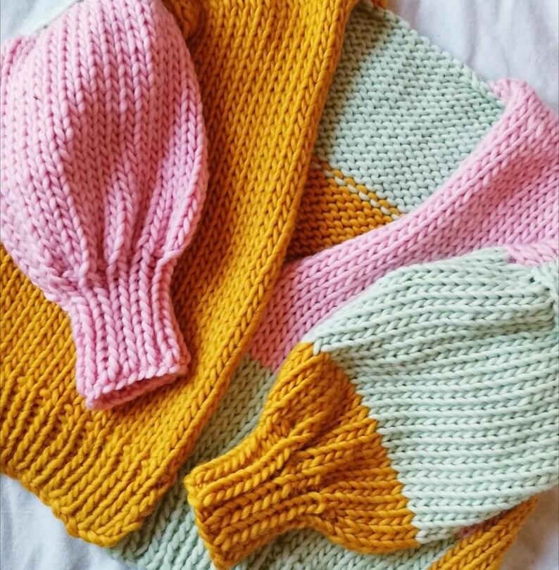 May include: A close-up view of a hand-knitted sweater in shades of pink, mustard yellow, and mint green. The sweater features large, puffy sleeves and a cozy, textured knit design. The color-blocked pattern adds a modern touch to the handmade garment.