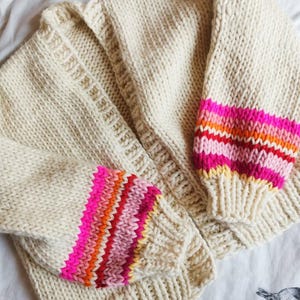 May include: Cream-colored, chunky knit cardigan with vibrant, striped sleeves. The sleeves feature bands of pink, orange, and red. The cardigan is a cozy, handmade garment, perfect for adding a pop of color to any outfit.