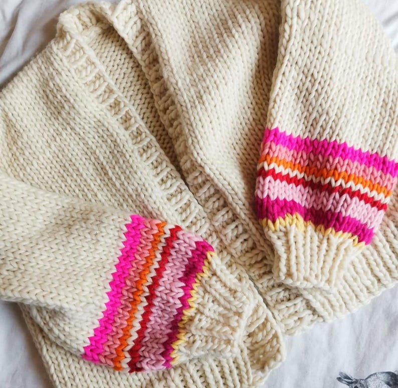 May include: Cream-colored, chunky knit cardigan with vibrant, striped sleeves. The sleeves feature bands of pink, orange, and red. The cardigan is a cozy, handmade garment, perfect for adding a pop of color to any outfit.