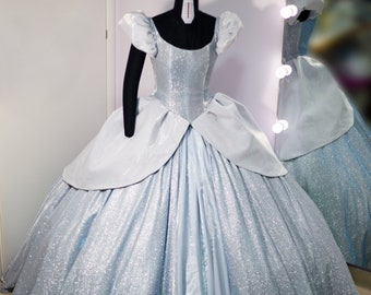May include: A light blue ball gown on a mannequin, featuring a scoop neckline, short puffed sleeves, and a voluminous skirt with a shimmering, glittery finish. The waist is accented with a structured peplum overlay, and the bodice is fitted.