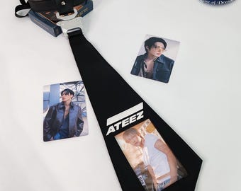 May include: A black necktie featuring a silver-toned metal buckle at the top, the white text ATEEZ printed in the center, and a clear plastic pocket displaying a photograph of a person. Two additional rectangular photo cards are included.
