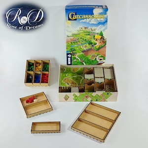 May include: A Carcassonne board game box with a colourful illustrated cover featuring a medieval landscape, accompanied by several light-coloured wooden storage organisers with various compartments for game pieces, including small red and white tokens.