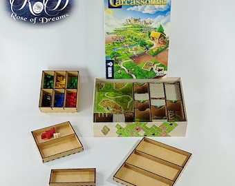 May include: A Carcassonne board game box with a colorful illustrated cover featuring a medieval landscape, accompanied by several light-colored wooden storage organizers with various compartments for game pieces, including small red and white tokens.
