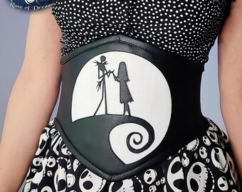 May include: Black corset belt featuring a white silhouette of Jack and Sally from The Nightmare Before Christmas against a full moon. The belt has a curved design and a spiral detail. The model is wearing a black and white polka dot top and a skirt with skull print.