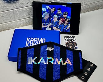 May include: A black and blue striped belt featuring the word KARMA in iridescent lettering and SKZ in white text. The set includes a blue box with the word KARMA, a tablet displaying a group of people, and a small card with a group photograph.