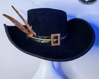May include: A black wide-brimmed hat with a flat crown, featuring a textured black band adorned with a rectangular gold-toned buckle and two brown and black patterned feathers tucked into the side. The hat has a braided edge along the brim.