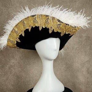 May include: A black hat with gold lace trim and white feathers. The hat has a wide brim and is displayed on a white mannequin head. The gold trim is intricately designed, adding a touch of elegance to the hat's overall appearance. The white feathers are placed around the hat.