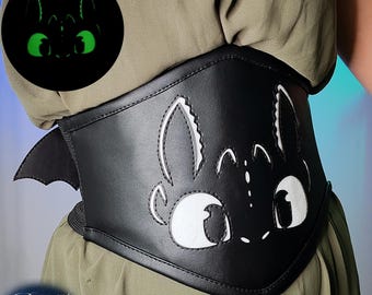 May include: A black faux leather corset belt featuring a white stitched graphic of a dragon face with large eyes and pointed ears. The design includes a small wing detail on the side and a circular inset showing the graphic glowing in the dark.