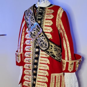 May include: A red and gold historical costume with a black hat and white feather trim. The jacket features gold trim, buttons, and a decorative sash. The costume is displayed on a mannequin.