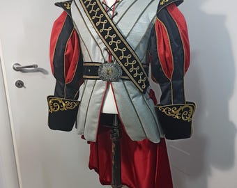 May include: A silver and black costume jacket with red accents and gold embroidery. The jacket has a high collar and a long, flowing tail. The jacket is worn over a white shirt with a black lace-up collar. The jacket is worn with a black belt with a gold buckle and a gold sash with black embroidery.