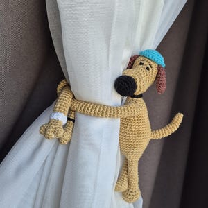 以下が含まれることがあります： ベージュ色の犬の編みぐるみカーテンタッセル。黒い鼻、青い帽子、茶色の耳が特徴です。犬は白いカーテンを抱きしめており、背景には茶色のカーテンがあります。犬の腕と脚はカーテンに巻き付いています。