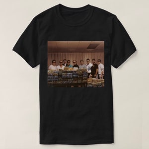 May include: A black and white photograph of a group of people standing in front of a large stack of cash. The people are wearing dark clothing and have their faces obscured. The cash is stacked in neat rows and is likely US dollars.