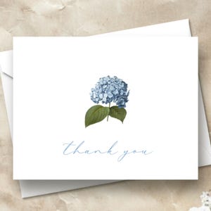 May include: White thank you card with a blue hydrangea flower illustration and the words "thank you" in a delicate script. The card is placed on a textured, light brown surface, with a white envelope and sprigs of white flowers.
