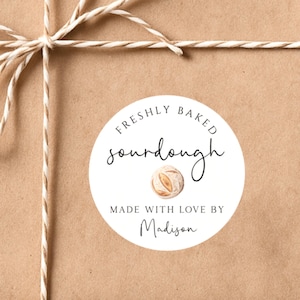 May include: A brown paper package tied with twine, featuring a white circular label. The label reads "FRESHLY BAKED sourdough MADE WITH LOVE BY Madison" in a mix of script and sans-serif fonts, with a watercolor illustration of bread.