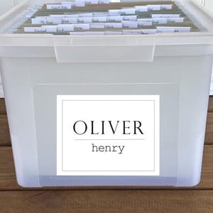 May include: A clear plastic storage bin with a lid, filled with labeled file folders. The bin has a white label with the name "OLIVER" and "henry" printed on it. The bin is sitting on a wooden surface.