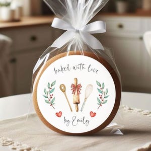May include: A round cookie in a clear plastic bag tied with a white ribbon. The cookie features a label with the text baked with love by Emily, illustrations of a wooden spoon, a rolling pin with a red plaid bow, a whisk, and two red hearts.