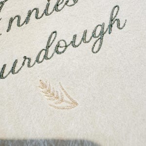 May include: An off-white fabric featuring dark green embroidered cursive text that reads "ennie" and "urdough" with a small, light beige embroidered wheat stalk design positioned below the text, showcasing detailed thread work on a plain textile.