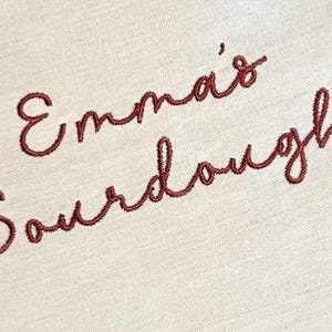 Puede incluir: Una tela de color beige claro con el texto bordado Emma's Sourdough en una escritura cursiva de color marr&oacute;n rojizo oscuro. El bordado est&aacute; centrado en el material tejido, mostrando un patr&oacute;n de puntadas limpio y detallado en la superficie.