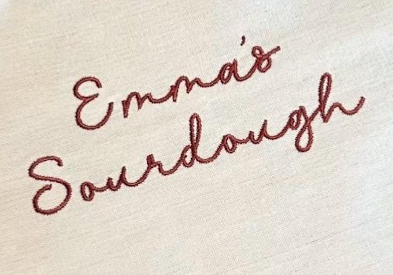 Puede incluir: Una tela de color beige claro con el texto bordado Emma's Sourdough en una escritura cursiva de color marr&oacute;n rojizo oscuro. El bordado est&aacute; centrado en el material tejido, mostrando un patr&oacute;n de puntadas limpio y detallado en la superficie.