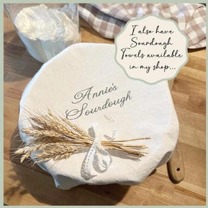 May include: A white sourdough bread cover with the text "Annie's Sourdough" and a wheat bundle tied with lace. A bag of flour is in the background. The image also includes the text "I also have Sourdough Towels available in my shop..."