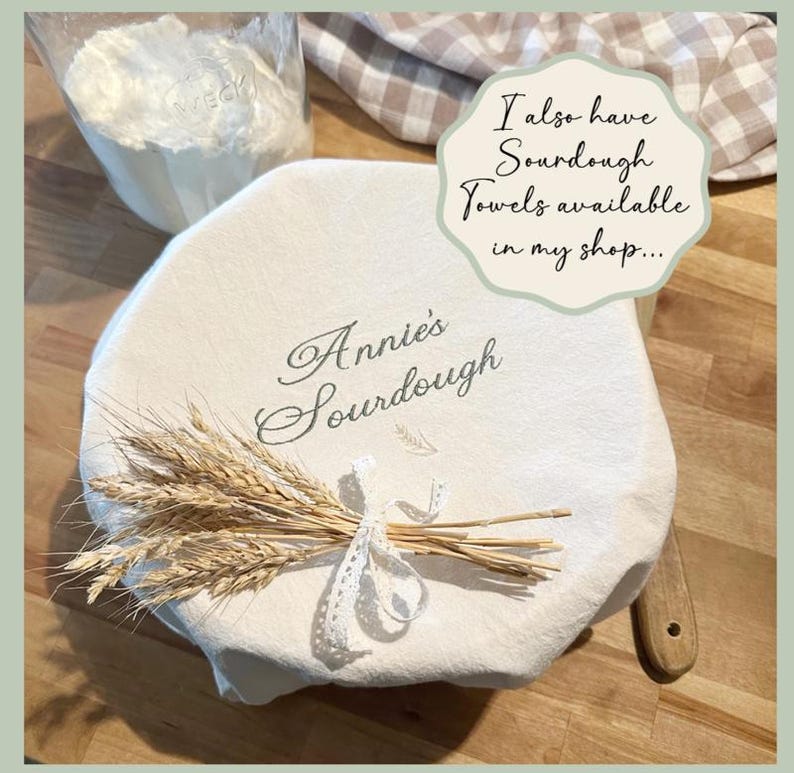 May include: A white sourdough bread cover with the text "Annie's Sourdough" and a wheat bundle tied with lace. A bag of flour is in the background. The image also includes the text "I also have Sourdough Towels available in my shop..."