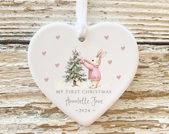 Könnte beinhalten: Weißer herzförmiger Keramik-Anhänger mit einem rosa Hasen, der einen Weihnachtsbaum hält. Der Anhänger ist mit rosa Herzen und dem Text "My First Christmas Annabelle Jane - 2024 - " verziert.