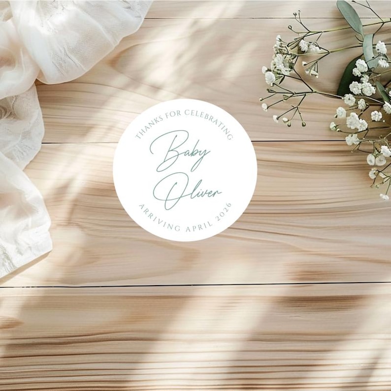 Puede incluir: Una pegatina redonda blanca con el texto "THANKS FOR CELEBRATING Baby Oliver ARRIVING APRIL 2026" en escritura turquesa. La pegatina est&aacute; sobre una superficie de madera marr&oacute;n claro, con flores blancas y tela en el fondo.